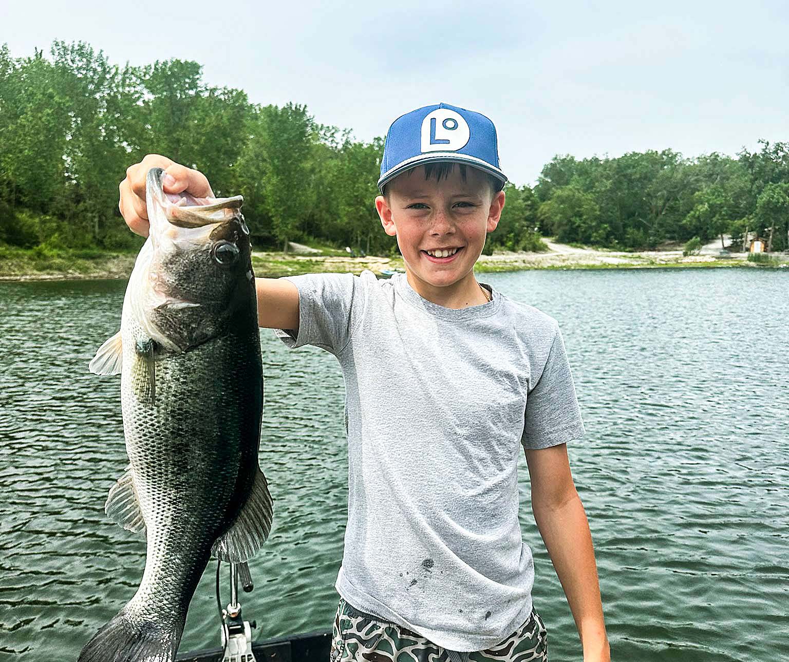 A boy in a Landoption cap holding a largemouth bass by a lake